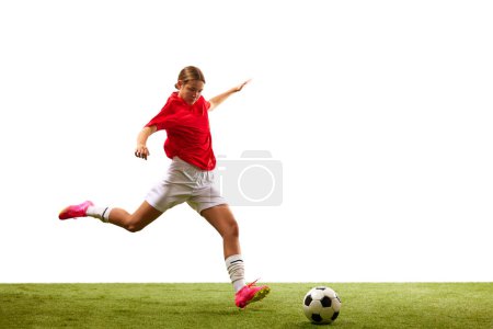 Téléchargez les photos : Jeune fille concentrée, footballeuse en mouvement, jouant sur l'herbe verte, prête à frapper balle isolée sur fond blanc. Concept de sport, compétition, action, succès, vainqueur. Espace de copie pour la publicité - en image libre de droit