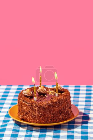 Téléchargez les photos : Gâteau délicieux au chocolat sur assiette avec des bougies sur nappe à carreaux bleus sur fond rose. Concept de nourriture, desserts, fête d'anniversaire, fête. Style pop art. Espace de copie pour l'annonce. Affiche - en image libre de droit