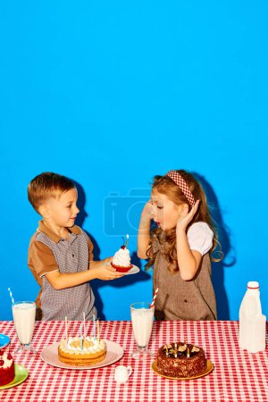 Photo for Little boy, brother giving birthday cake with candle to his sister against blue studio background. Siblings birthday. Concept of childhood, birthday celebration, family, fun, food - Royalty Free Image