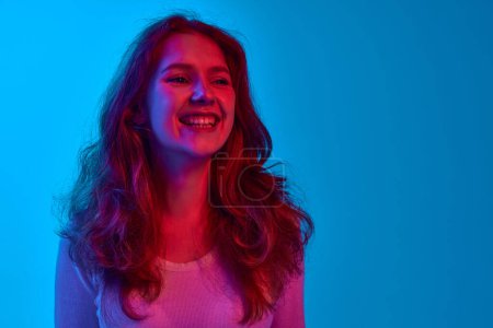 Photo for Portrait of beautiful, happy, cheerful young girl with wavy hair smiling against blue studio background in neon light. Concept of youth, human emotions, facial expression, lifestyle - Royalty Free Image