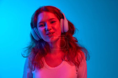 Photo for Close-up of young girl listening to music in headphones and singing against blue studio background in neon light. Concept of youth, human emotions, facial expression, lifestyle - Royalty Free Image