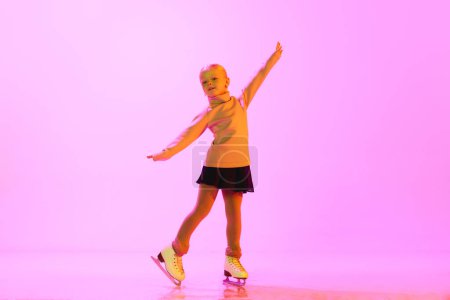 Téléchargez les photos : Petite, belle, mignonne fille de 5 ans en pull et jupe debout sur des patins, s'entraînant sur fond rose au néon. Concept d'enfance, patinage artistique sport, passe-temps, école - en image libre de droit