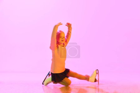 Foto de Hermosa, linda niña, niño, entrenamiento patinaje artístico sobre fondo rosa en luz de neón. Niño en suéter y falda. Concepto de infancia, patinaje artístico deporte, hobby, escuela, educación - Imagen libre de derechos