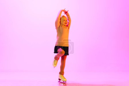 Foto de Sonriente, feliz, niño pequeño positivo, niña en suéter lindo y falda, bailando en patines sobre fondo rosa en neón. Concepto de infancia, patinaje artístico deporte, hobby, escuela, educación - Imagen libre de derechos