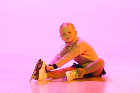 Foto de Niña adorable, chide en suéter y falda, sentada en el suelo y atando cordones en patines sobre fondo rosa en neón. Concepto de infancia, patinaje artístico deporte, hobby, escuela, educación - Imagen libre de derechos