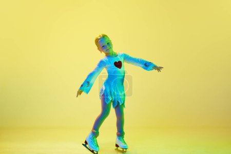 Photo for Full-length of cute, beautiful adorable kid, little girl, dancing, performing figure skating against yellow background in neon. Concept of childhood, figure skating sport, hobby, school, education - Royalty Free Image