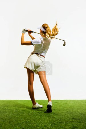 Photo for Back view of slim, blonde, young girl in stylish white clothes playing golf on grass, teeing off isolated over white background. Concept of sport, hobby, beauty and fashion, relaxation, game - Royalty Free Image