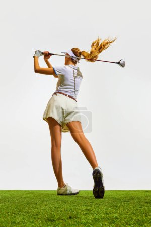 Photo for Beautiful, sportive, blonde, young girl with slim, fit body playing golf on grass, teeing off isolated over white background. Concept of sport, hobby, beauty and fashion, relaxation, game - Royalty Free Image