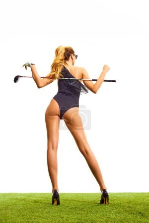 Foto de Mujer joven atractiva con cuerpo delgado y hermoso en un elegante traje de cuerpo posando sobre tacones con palo de golf aislado sobre fondo blanco. Vista trasera. Concepto de deporte, hobby, belleza y moda, relajación, juego - Imagen libre de derechos