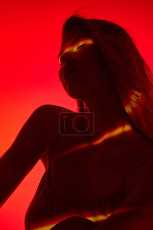 Foto de Mujer joven sensual y tierna en la parte superior posando con los ojos vestidos sobre fondo rojo con reflexión de neón en el cuerpo. Concepto de arte moderno, belleza, estilo, futurismo y cyberpunk, creatividad - Imagen libre de derechos