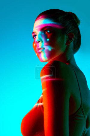 Téléchargez les photos : Portrait de belle jeune femme sur fond bleu avec réflexion néon multicolore sur le visage. Regarde bien. Concept d'art moderne, beauté, style, futurisme et cyberpunk, créativité - en image libre de droit