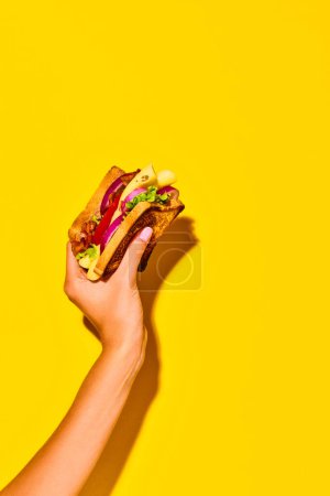 Photo for Woman holding delicious sandwich with grilled bread, ham, lettuce, cheese, tomato, onion and avocado over yellow background. Concept of food, taste, breakfast. Complementary colors. Poster, ad - Royalty Free Image
