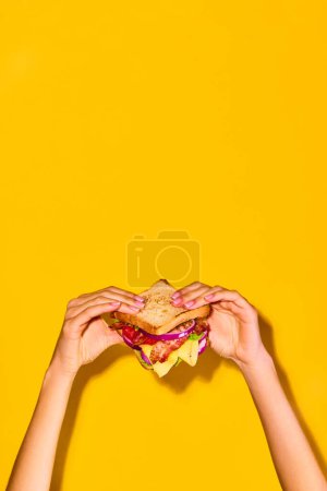 Photo for Woman holding delicious sandwich with grilled bread, ham, lettuce, cheese, tomato, onion and avocado over yellow background. Concept of food, taste, breakfast. Complementary colors. Poster, ad - Royalty Free Image