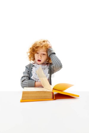 Photo for Little redhead, curly girl sitting at desk put her hand on head while reading book. Child study and learn for preschool. Online education. Concept of childhood, International Knowledge Day. Copy space - Royalty Free Image