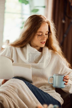 Photo for Portrait of young attractive girl in weights who reads book and drinks tea while sitting at home on the sofa after hard working week. concept of lifestyle, winter holiday season, weekend, relax, cozy - Royalty Free Image