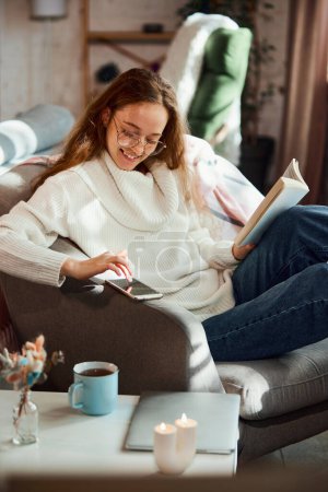 Photo for Beautiful young girl in glasses and knitted warm sweater reading book and answer to incoming messages. concept of lifestyle, winter holiday season, autumn weekend, relax and cozy atmosphere, - Royalty Free Image