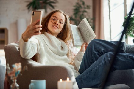 Téléchargez les photos : Jeune fille attrayante faire selfie tout en lisant le livre à la maison atmosphère confortable et relaxante. Concept de style de vie, vacances d'hiver, week-end d'automne, détente et atmosphère chaleureuse, - en image libre de droit