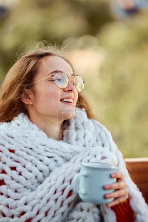 Photo for Portrait of happy smiling young girl sits covered blanket and drinks warming tea on bench in park. Autumn mood. concept of lifestyle, winter holiday season, weekend, relax and cozy atmosphere, - Royalty Free Image
