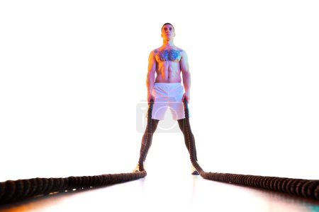 Photo for Muscular young man with fit, strong shirtless body standing with battle rope against white studio background in neon light. Concept of sport, active and healthy lifestyle, body care. Copy space for ad - Royalty Free Image