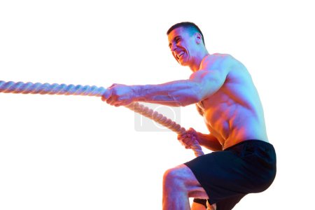 Photo for Shirtless young man with strong body and hands pulling rope, training against white studio background in neon light. Concept of sport, active and healthy lifestyle, body care, fitness - Royalty Free Image