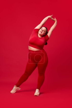 Foto de Hermosa mujer con sobrepeso en el entrenamiento de ropa deportiva cómoda, que se extiende contra el fondo rojo del estudio. Concepto de deporte, positividad corporal, pérdida de peso, cuidado del cuerpo y de la salud, autoaceptación - Imagen libre de derechos