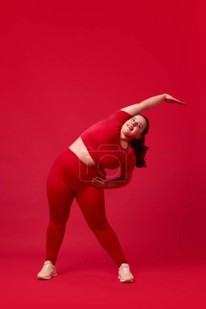 Foto de Mujer joven con sobrepeso en entrenamiento de ropa deportiva, haciendo ejercicios, estirándose contra el fondo rojo del estudio. Modelo de talla grande. Concepto de deporte, positividad corporal, pérdida de peso, cuidado corporal y de la salud - Imagen libre de derechos