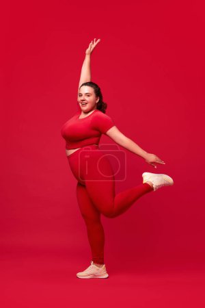 Foto de Mujer joven feliz, sonriente y hermosa con entrenamiento en forma de cuerpo con sobrepeso sobre fondo de estudio rojo. Concepto de deporte, positividad corporal, pérdida de peso, cuidado corporal y de la salud - Imagen libre de derechos