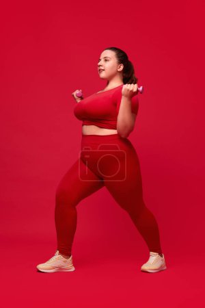 Foto de Sobrepeso joven hermosa mujer de entrenamiento, haciendo ejercicios con mancuernas contra fondo de estudio rojo. Concepto de deporte, positividad corporal, pérdida de peso, cuidado corporal y de la salud - Imagen libre de derechos