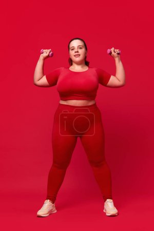 Foto de Sobrepeso joven hermosa mujer de entrenamiento, haciendo ejercicios con mancuernas contra fondo de estudio rojo. Concepto de deporte, positividad corporal, pérdida de peso, cuidado corporal y de la salud - Imagen libre de derechos