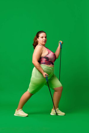 Photo for Motivated young overweigh woman in sportswear training, doing exercises with fitness expanders against green studio background. Concept of sport, body-positivity, weight loss, body and health care - Royalty Free Image