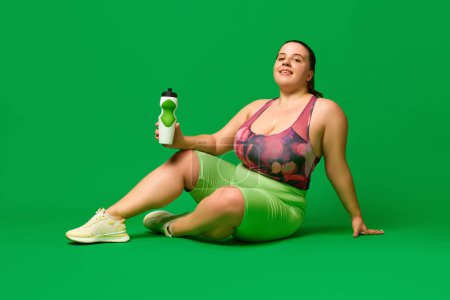 Foto de Hidratación. Modelo de talla grande, mujer joven con sobrepeso en ropa deportiva sentada en el suelo con botella de agua sobre fondo de estudio verde. Concepto de deporte, positividad corporal, pérdida de peso, cuerpo, atención médica - Imagen libre de derechos