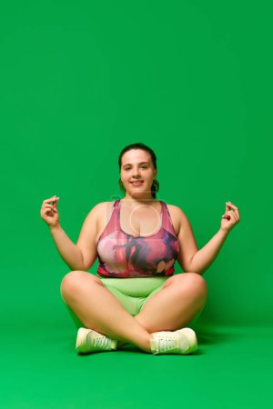 Téléchargez les photos : Jeune belle femme de surpoids assis sur la pose de lotus, formation, ding yoga sur fond de studio vert. Calme l'esprit et le corps. Concept de sport, positivité corporelle, perte de poids, corps et soins de santé - en image libre de droit