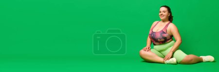 Foto de Modelo de talla grande, mujer joven con sobrepeso en ropa deportiva sentada en el suelo contra el fondo verde del estudio. Concepto de deporte, positividad corporal, pérdida de peso, cuidado corporal y de la salud. Banner - Imagen libre de derechos