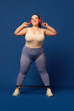 Photo for Young overweigh woman in headphones training with fitness elastic band s against blue background. against blue studio background. Concept of sport, body-positivity, weight loss, body and health care. - Royalty Free Image