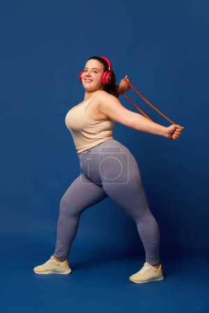 Photo for Smiling young woman with fat body training with elastic bands, fitness expanders against blue studio background. Concept of sport, body-positivity, weight loss, body and health care. Copy space for ad - Royalty Free Image
