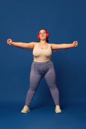 Photo for Overweigh young woman training with fitness expanders against blue studio background. Concept of sport, body-positivity, weight loss, body and health care. Copy space for ad - Royalty Free Image