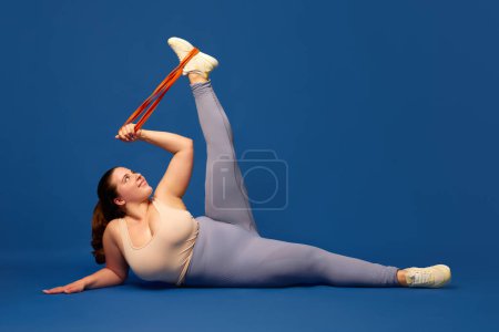 Photo for Leg swings. Young overweigh woman training with fitness elastic band against blue studio background. Concept of sport, body-positivity, weight loss, body and health care. Copy space for ad - Royalty Free Image
