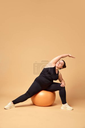 Photo for Stretching and balance. Young overweigh woman in black sportswear training with fitness ball against beige studio background. Concept of sport, body-positivity, weight loss, body and health care. - Royalty Free Image
