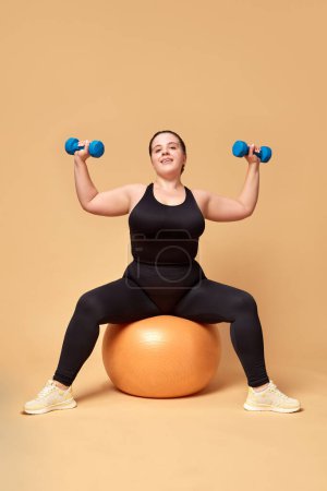 Photo for Young overweigh woman in black sportswear training with fitness ball and dumbbells against beige studio background. Concept of sport, body-positivity, weight loss, body, health care. Copy space for ad - Royalty Free Image