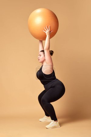 Photo for Young overweigh woman in black sportswear training with fitness ball, doing squats against beige studio background. Concept of sport, body-positivity, weight loss, body, health care. Copy space for ad - Royalty Free Image