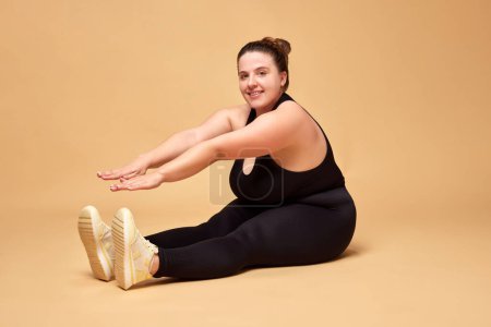 Photo for Young woman with fat, overweigh body in black sportswear sitting and doing stretching exercises against beige studio background. Concept of sport, body-positivity, weight loss, body and health care. - Royalty Free Image