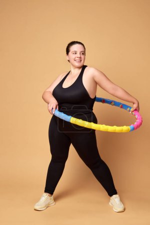 Foto de Mujer joven con sobrepeso en entrenamiento de ropa deportiva negra con aro contra fondo de estudio beige. Concepto de deporte, positividad corporal, pérdida de peso, cuidado corporal y de la salud. Copiar espacio para anuncio - Imagen libre de derechos