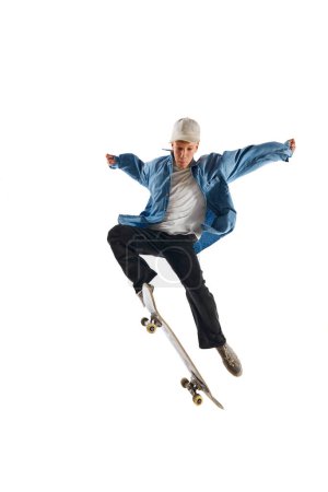 Téléchargez les photos : Jeune homme sportif et actif en tenue décontractée en mouvement, s'entraînant avec du skateboard, faisant des cascades isolées sur fond blanc. Concept de sport professionnel, compétition, entraînement, action. - en image libre de droit