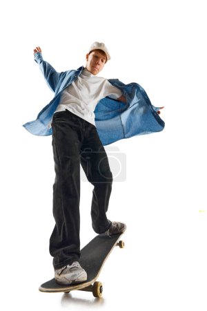 Foto de Niño adolescente con camisa vaquera azul y gorra en movimiento, entrenando con monopatín, haciendo acrobacias aisladas sobre fondo blanco. Concepto de deporte profesional, competición, entrenamiento, acción. Copiar espacio para anuncio - Imagen libre de derechos