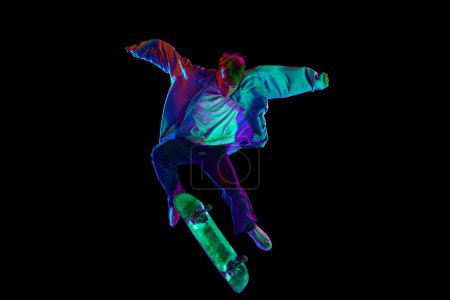 Photo for Teen boy, active guy in casual clothes in motion, training, practicing stunts on skateboard against black studio background in neon light. Concept of professional sport, competition, training, action - Royalty Free Image