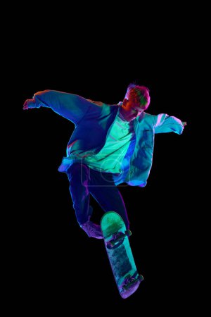 Photo for High jump. Active young man in casual clothes in motion, riding skateboard, training isolated against black studio background in neon. Concept of professional sport, competition, training, action - Royalty Free Image