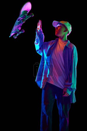 Photo for Boy in casual clothes standing with skateboard, posing, doing tricks against black studio background in neon light. Concept of professional sport, competition, training, action. Copy space for ad - Royalty Free Image