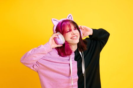 Photo for Happy, smiling young girl with piercing and two colored hair listening to music in headphones over vivid yellow studio background. Concept of youth, self-expression, fashion, emotions, positive mood - Royalty Free Image