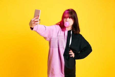 Foto de Chica joven con el pelo rosa en ropa casual tomando selfie con goma de mascar contra fondo de estudio amarillo vivo. Publicación en redes sociales. Concepto de juventud, auto-expresión, moda, emociones - Imagen libre de derechos