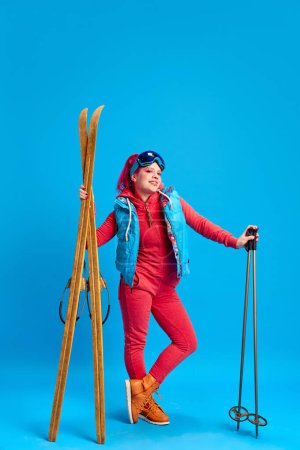 Photo for Young girl in colorful winter clothes standing with skis against blue studio background. Active holidays. Concept of youth, self-expression, winter vacation, active lifestyle, emotions - Royalty Free Image